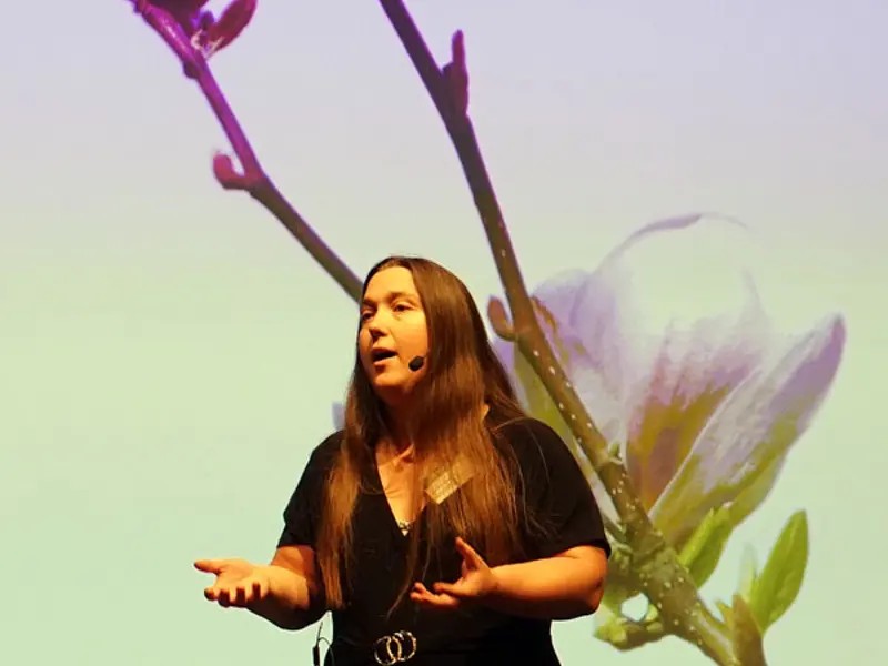 Woman talking in front of a picture of a flower branch.
