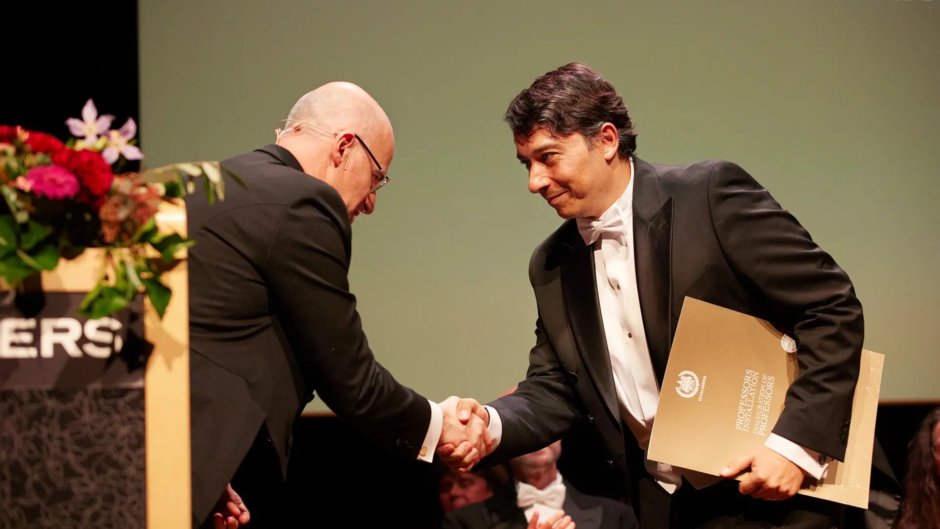 Two men shake hands with each other. One holds a diploma in his hand.