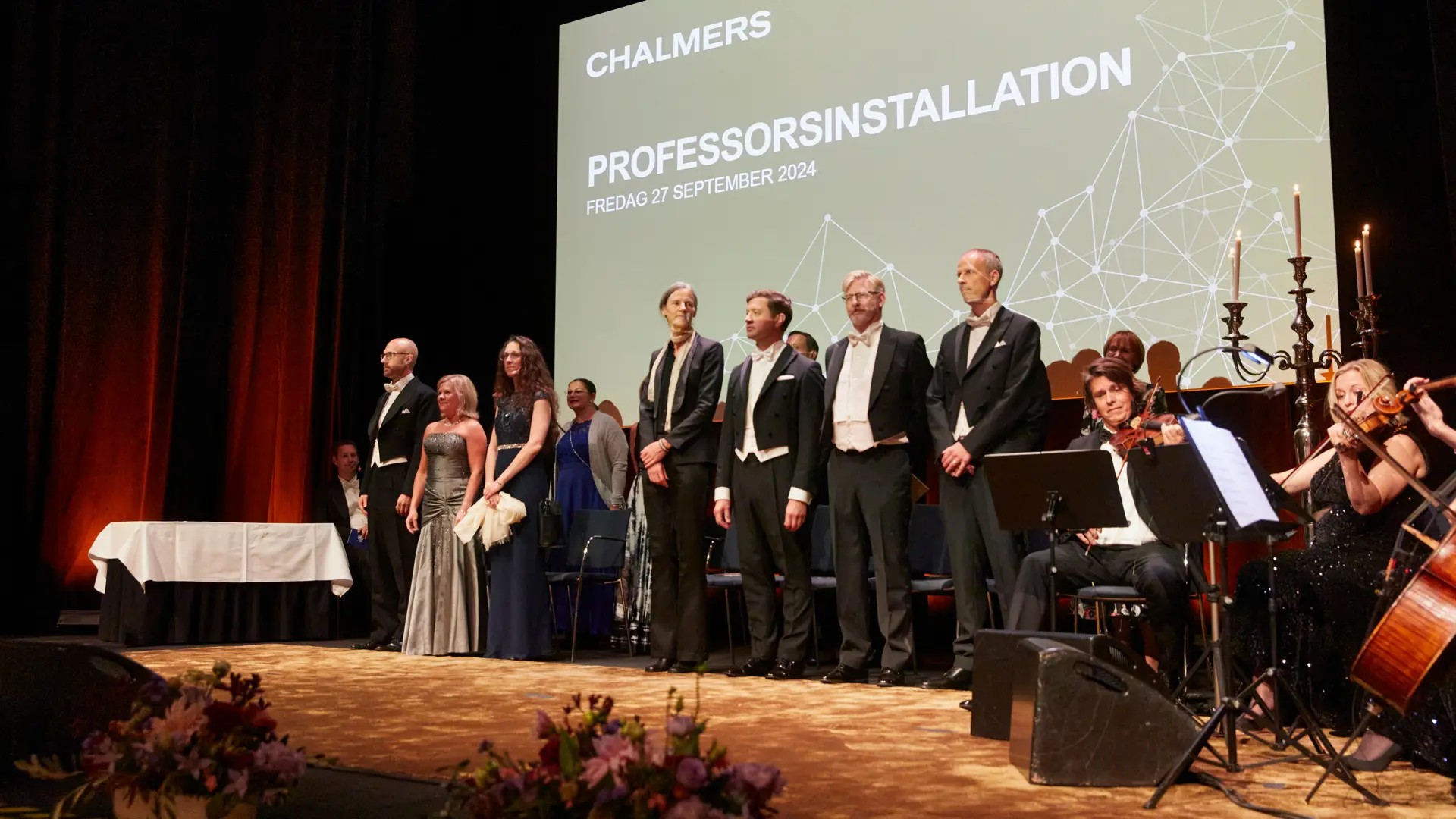 People in festive dress on a stage. Next to it, a violin quartet is playing.