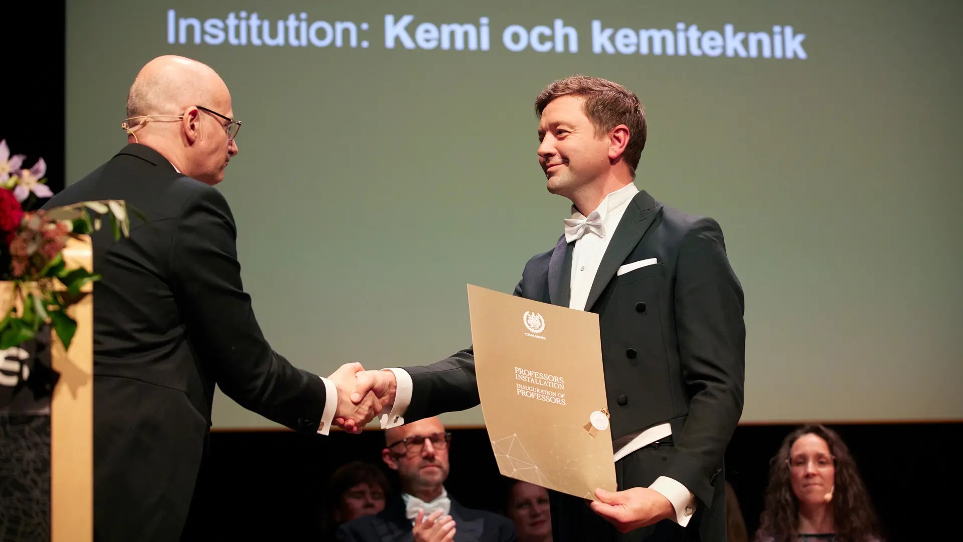 Two men shake hands with each other. One holds a diploma in his hand.