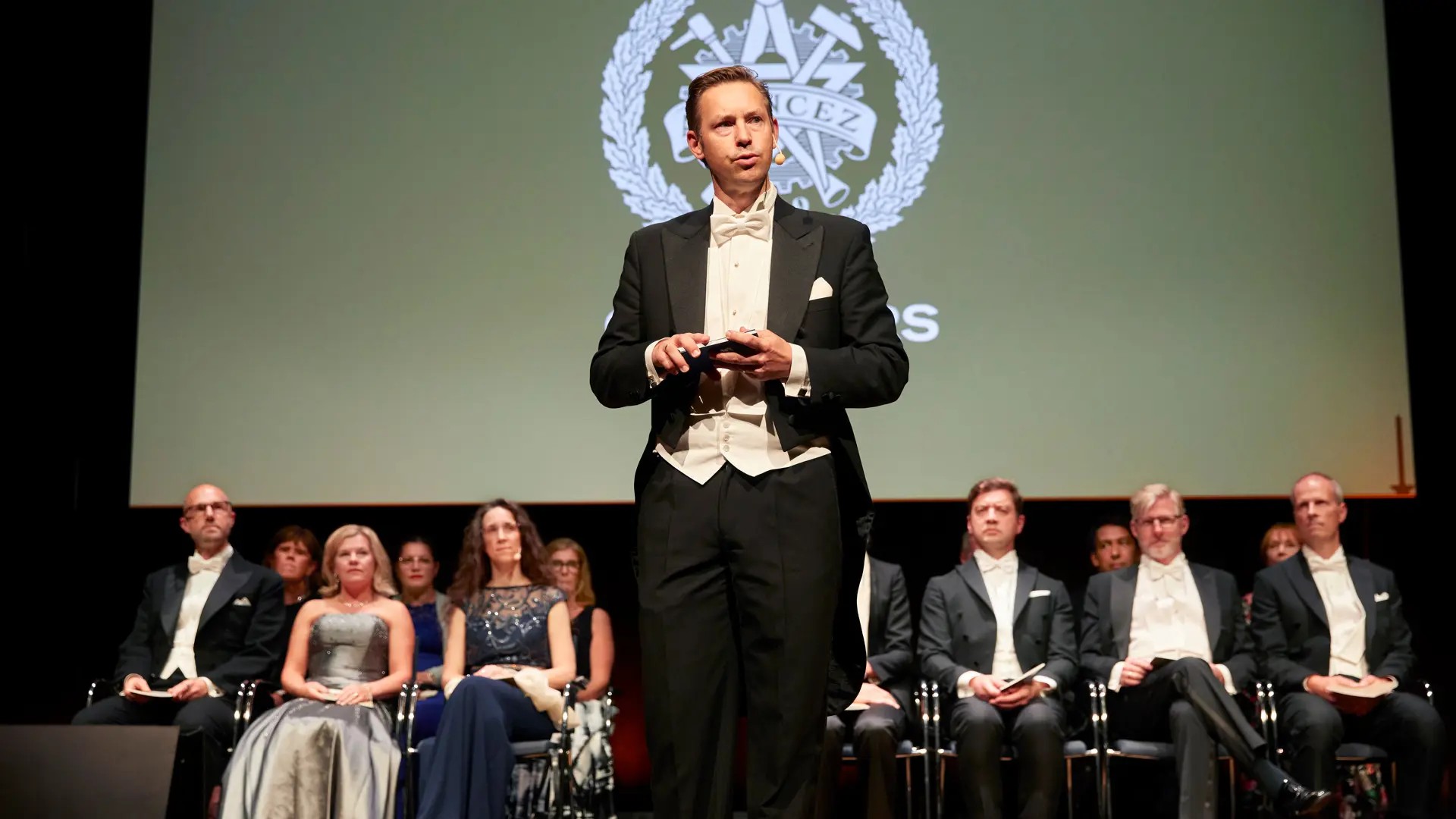 A man stands on stage in front of a row of seated professors.