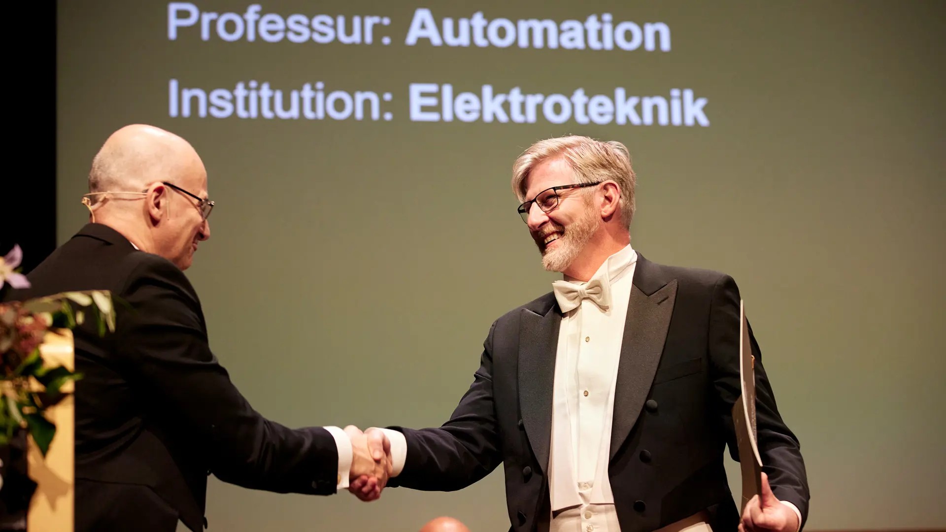 Two men shake hands with each other. One holds a diploma in his hand.